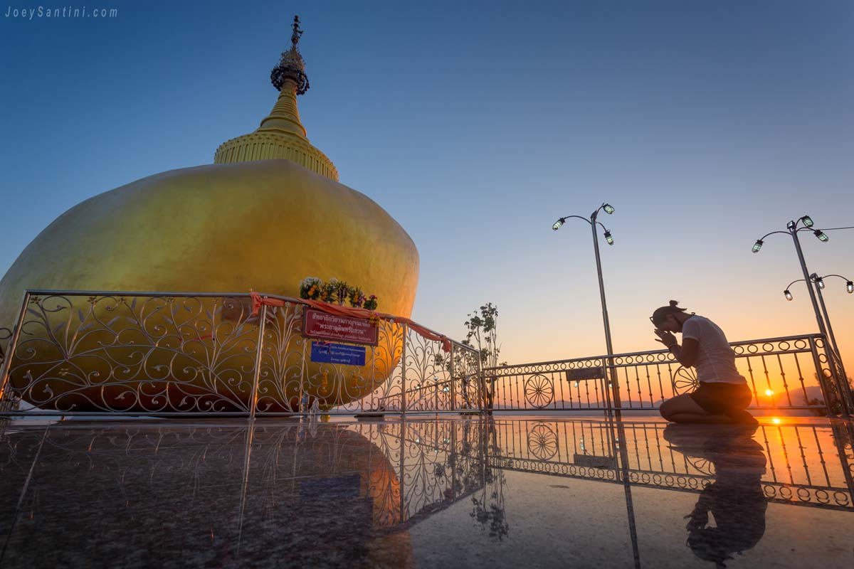 A worshipper kneeling in prayer beside the golden rock chedi at Wat Koh Siray during sunset