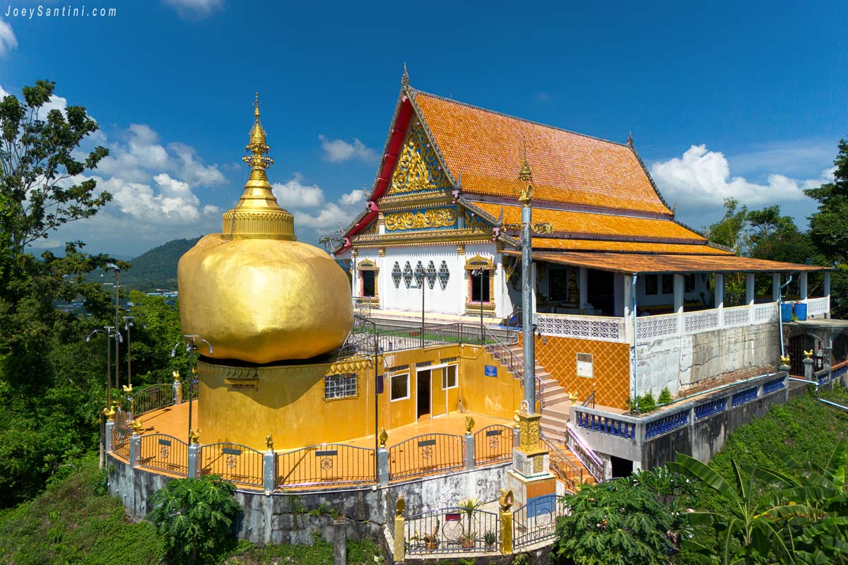 Overview of Wat Koh Siray in Phuket showing the large golden rock chedi and the traditional main temple building under a clear blue sky