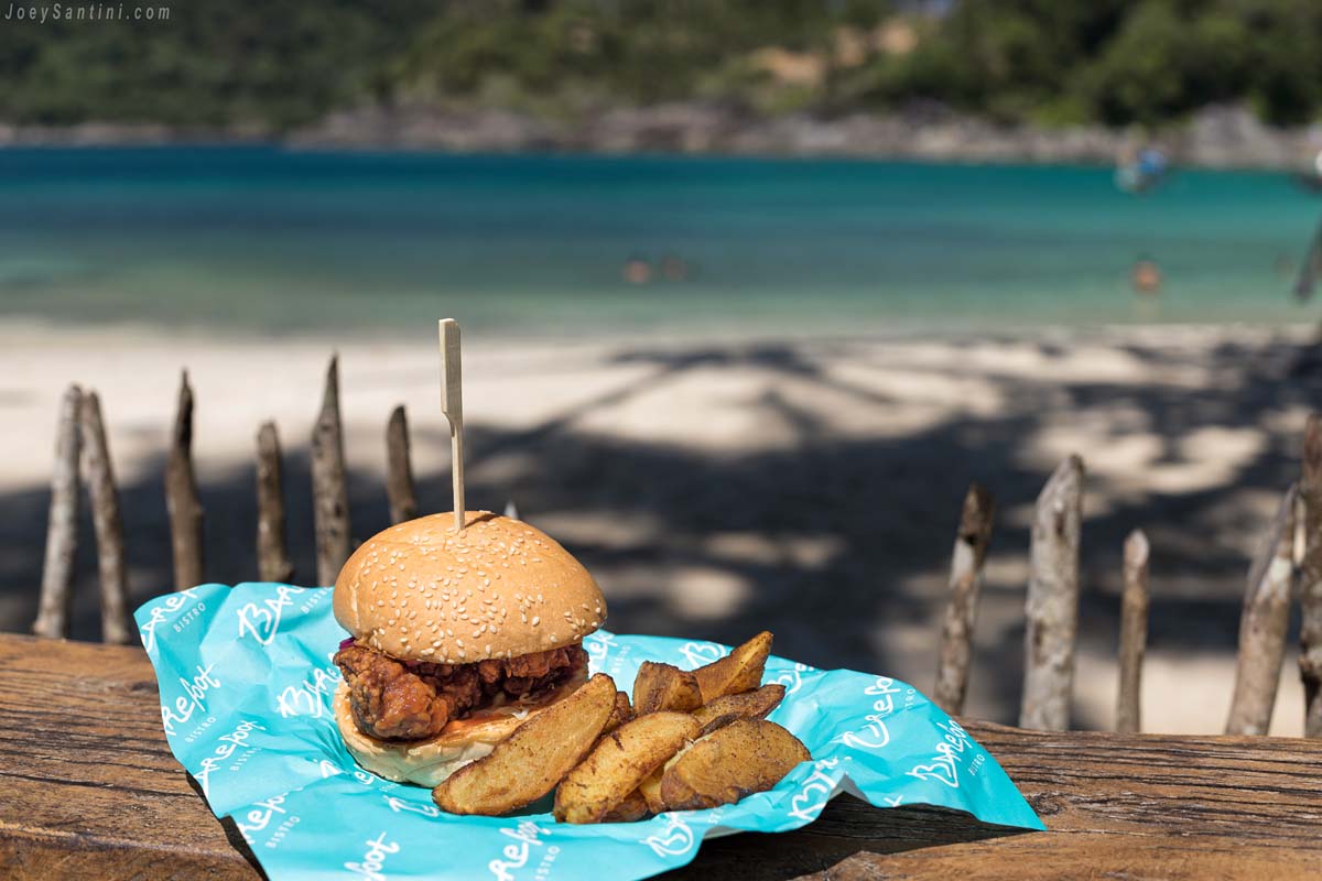 Crispy chicken burger served with potato wedges on branded paper at Barefoot Bistro with a tropical beach background.