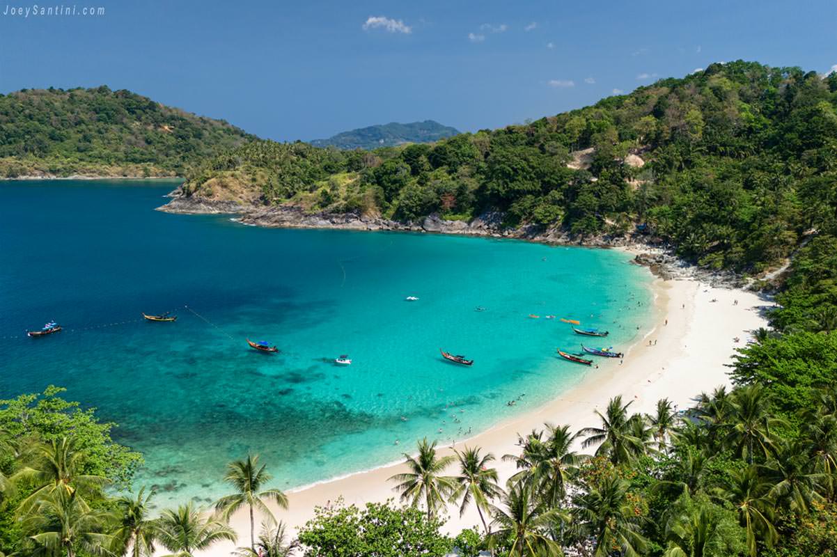 High-angle aerial view of Freedom Beach in Phuket, showing traditional Thai long-tail boats floating on crystal-clear turquoise water, surrounded by white sand and a dense tropical jungle.