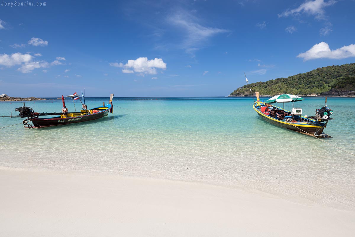 Two traditional Thai longtail boats anchored in the crystal-clear turquoise sea at Freedom Beach in Phuket under a bright blue sky.