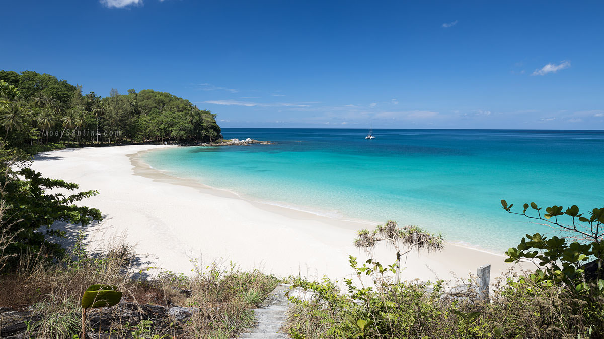 Panoramic viewpoint showing the pristine white sand, transparent turquoise water, and lush jungle headland of Freedom Beach in Phuket, as part of the Nak Kerd Protected Forest Area.