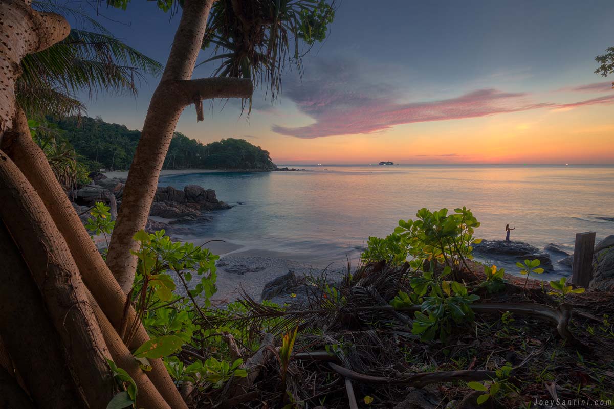 Dramatic sunset over Freedom Beach in Phuket with golden and pink sky reflections on the calm sea framed by coastal trees and rocks.