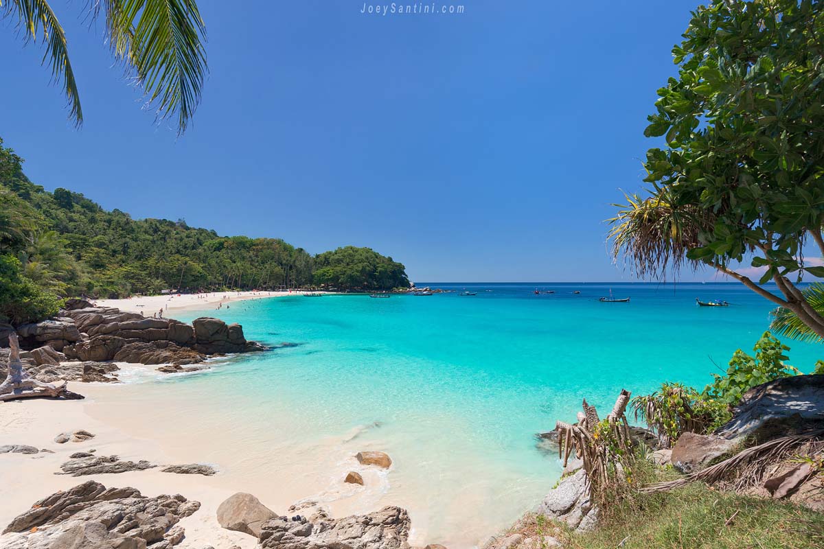 Panoramic view of Freedom Beach in Phuket featuring crystal-clear turquoise water, white sand beach, and traditional longtail boats under a clear blue sky.