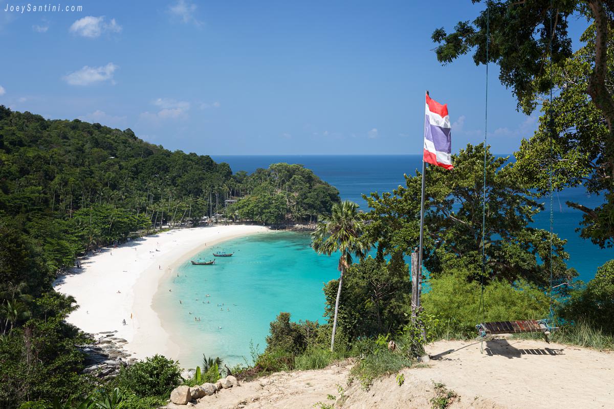 Stunning aerial view of Freedom Beach in Phuket, featuring a crescent of white sand, turquoise water, and a Thailand flag waving in the foreground.