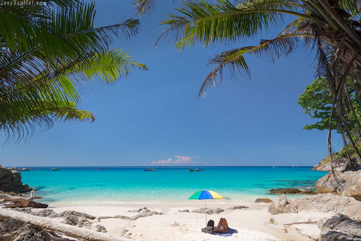 A person relaxing on a beach towel under a colorful beach umbrella on the white sand of Freedom Beach in Phuket with turquoise sea and palm trees.