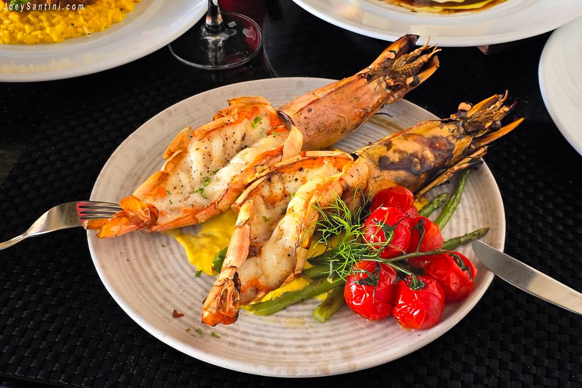 Grilled tiger prawns served on a black plate with fresh lime, red chili, and local green herbs at La Gritta restaurant.