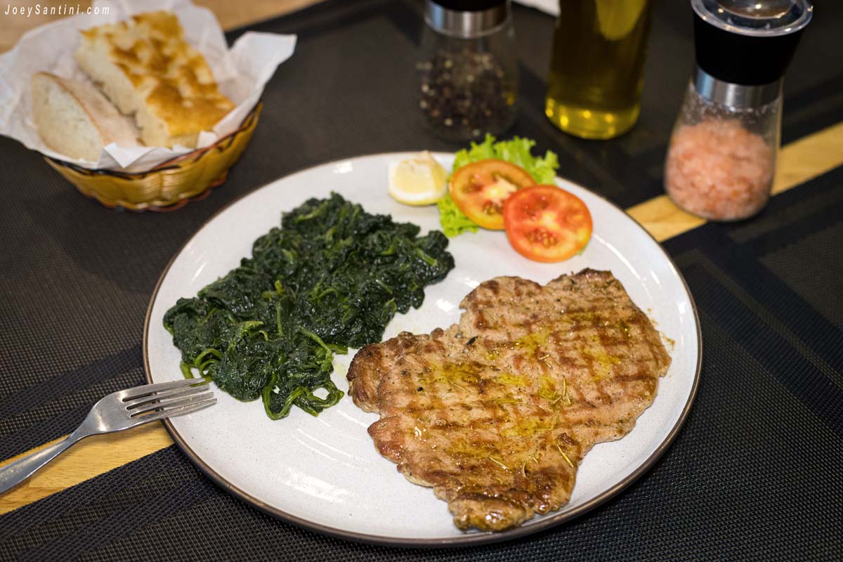 A plate of grilled beef paillard served with a side of sauteed spinach, sliced tomatoes, and lemon at Baia del Sole in Patong.
