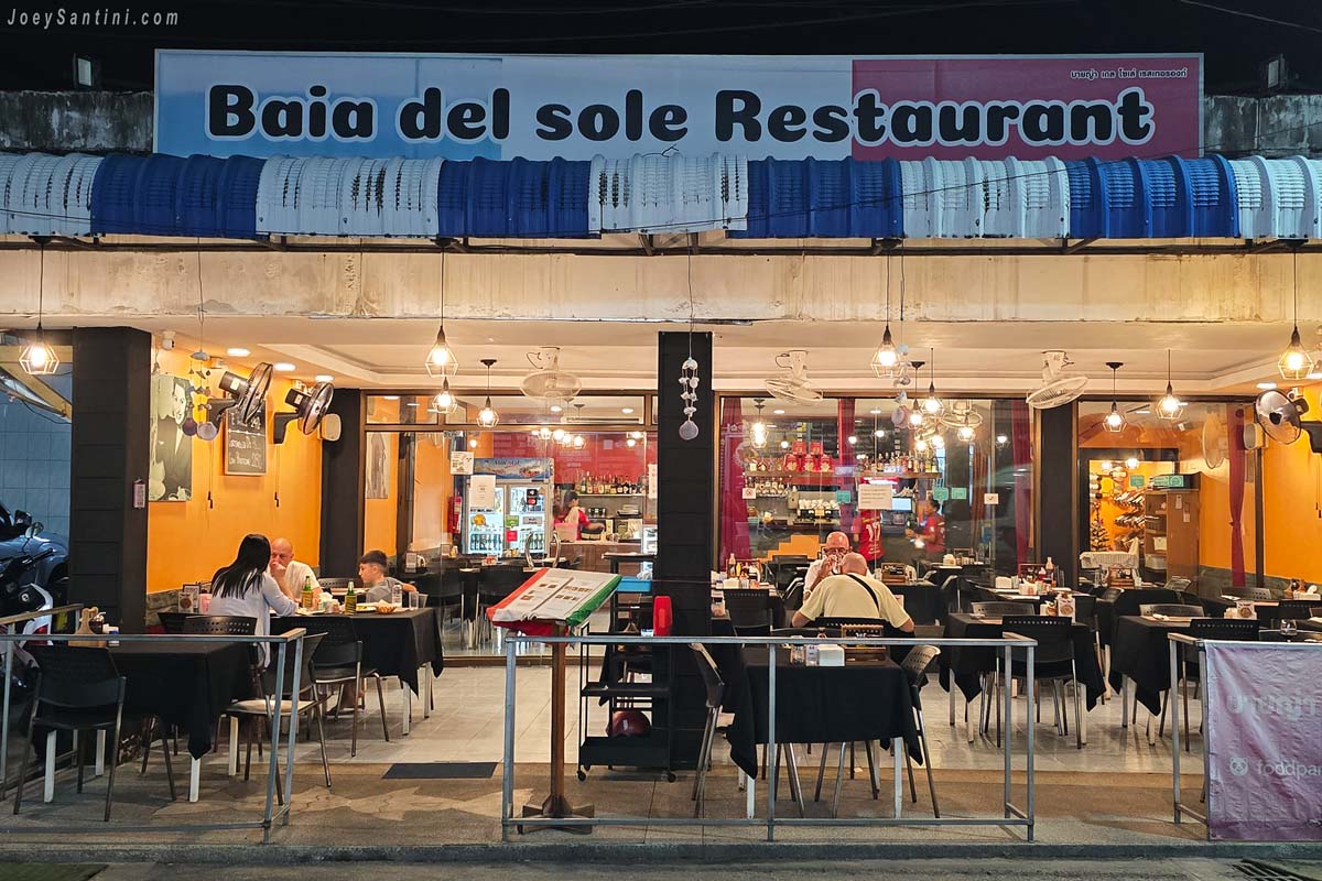 The illuminated exterior facade of Baia del Sole Italian restaurant in Patong at night, showing customers dining at tables under the main signboard.