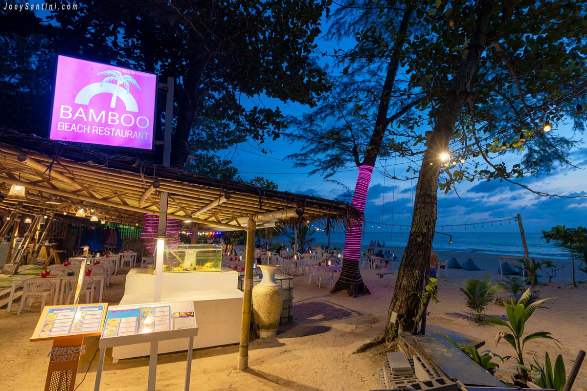 Bamboo Beach Restaurant illuminated with pink neon sign and warm hanging lights at night on Naithon Beach sand