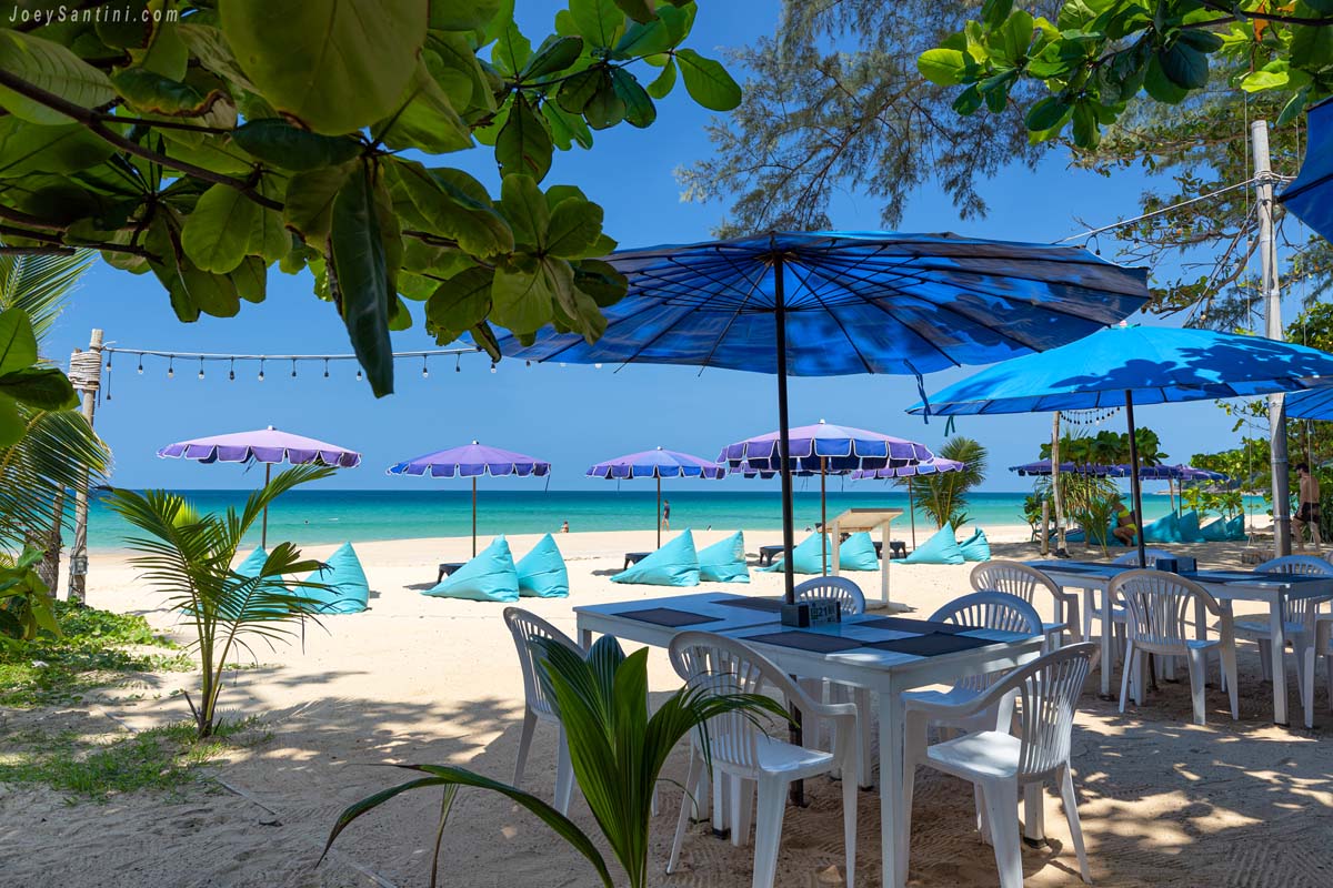 White dining tables blue umbrellas and turquoise bean bags on the white sand at Bamboo Beach Restaurant