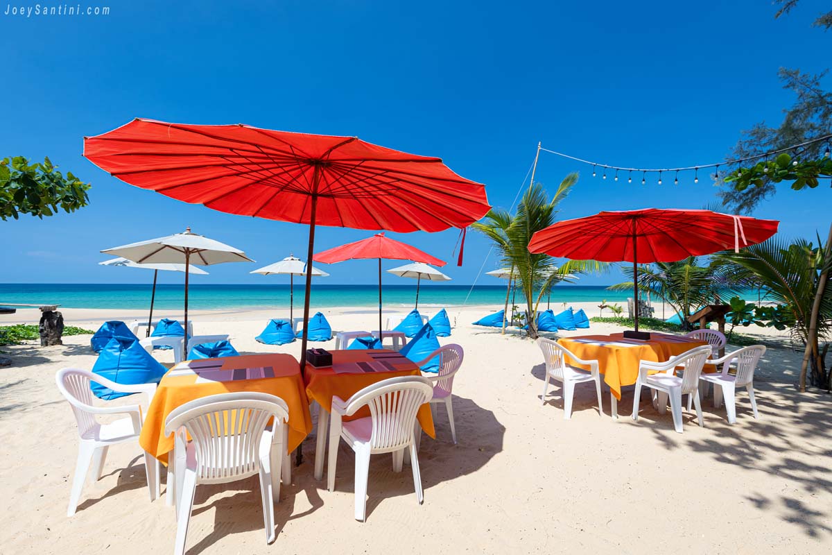 Inthanin Restaurant dining area on the white sand of Naithon Beach in Phuket with bright red umbrellas and blue bean bags