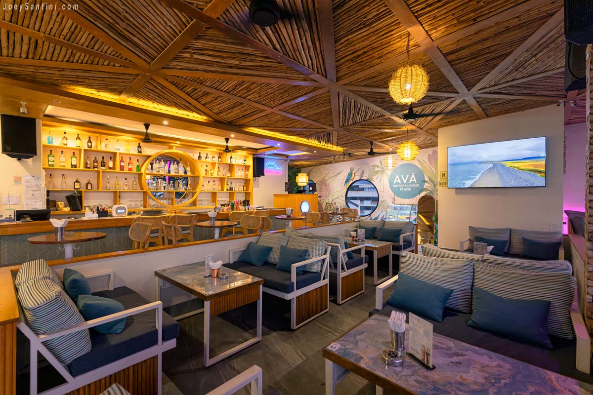 Indoor lounge area at AVA Rooftop Patong showing stylish blue and white striped cushions, wooden coffee tables, a fully-stocked cocktail bar, and the official brand logo on the wall.