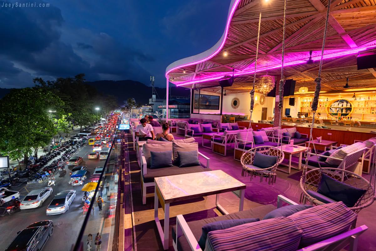 Cozy rooftop bar seating at AVA Rooftop Patong with wooden high chairs, round tables, and plush sofas set against a backdrop of green Casuarina pine trees during the golden hour.