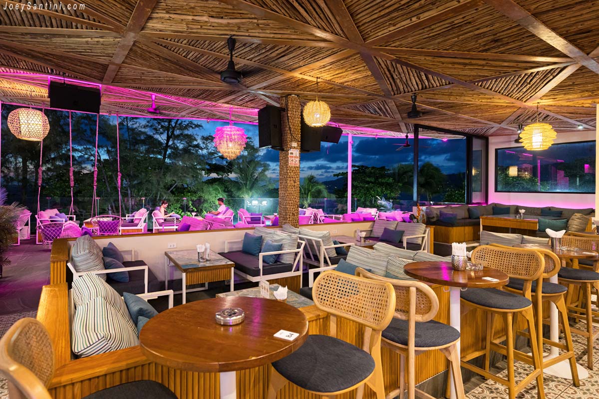 Cozy rooftop bar seating at AVA Rooftop Patong with wooden high chairs, round tables, and plush sofas set against a backdrop of green Casuarina pine trees during the golden hour.