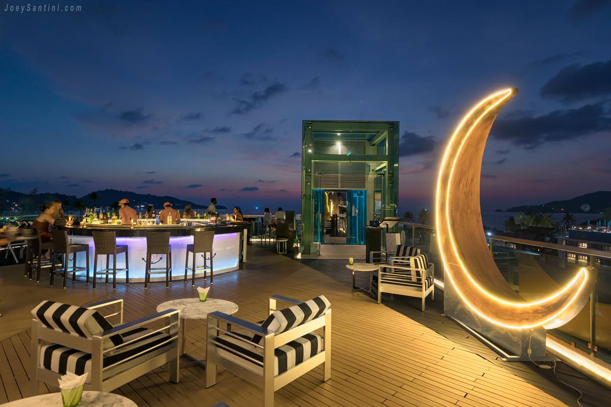 A large illuminated golden crescent moon decoration next to the outdoor lounge seating and circular bar at KEE Sky Lounge.