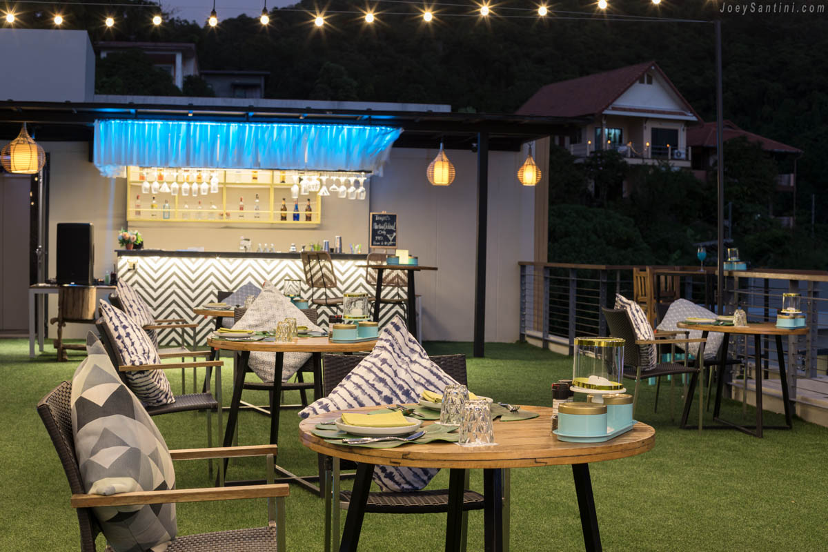 Table settings with patterned cushions in the foreground and an illuminated cocktail bar counter at Mojjo Rooftop Bar.