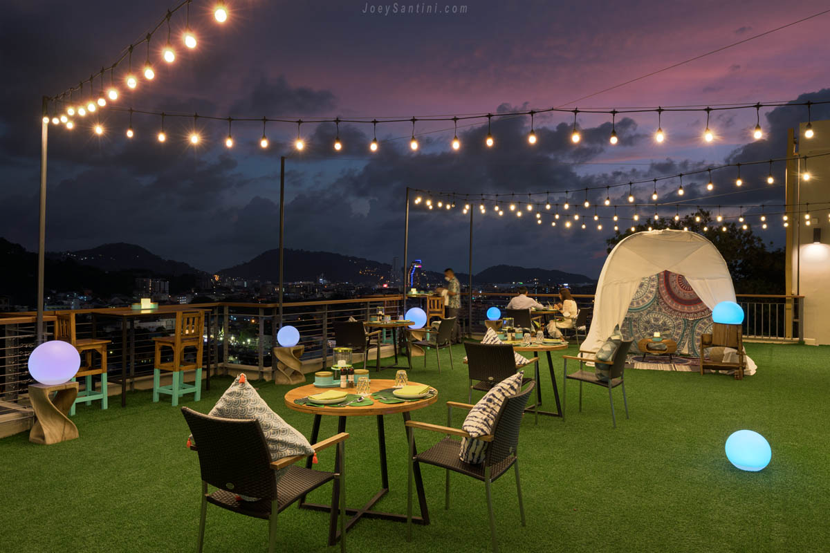 Outdoor rooftop garden terrace at Mojjo Rooftop Bar with artificial grass, hanging string lights, and glowing orb lamps under a purple dusk sky.