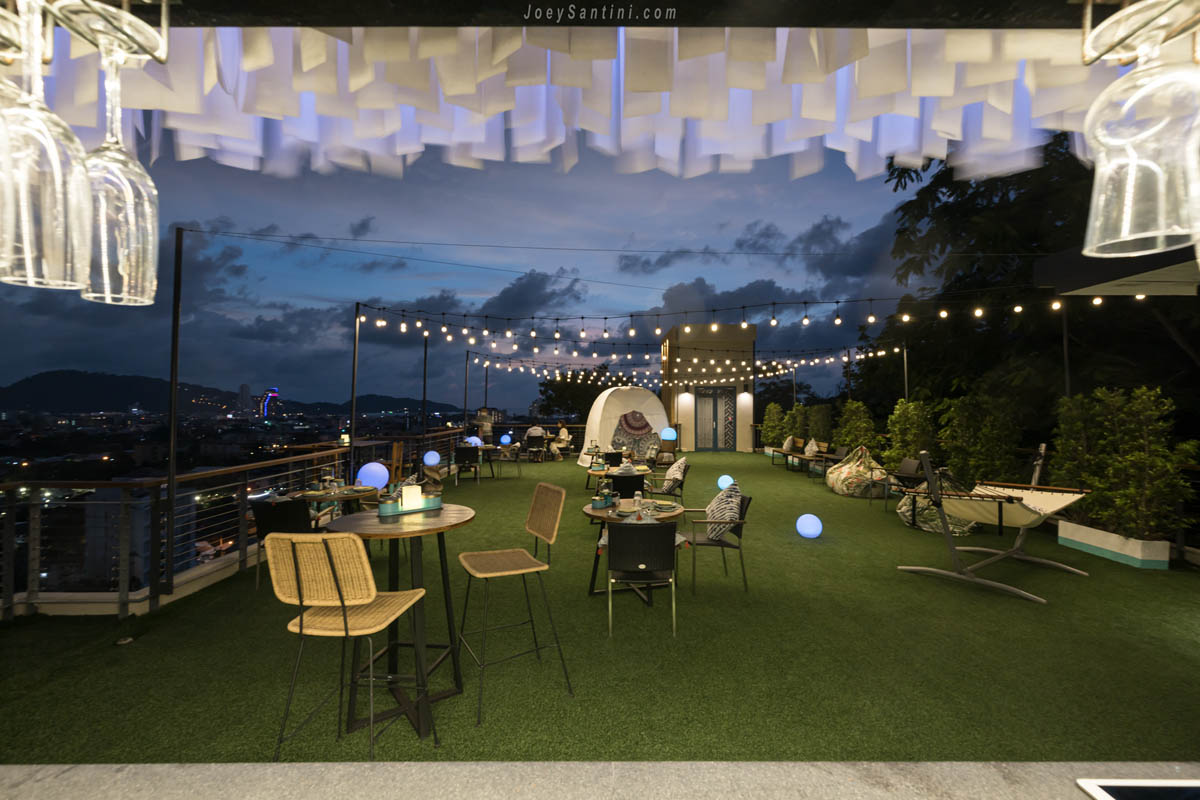 Wide view of Mojjo Rooftop Bar terrace from the bar counter featuring hammocks, bean bags, and hanging lights at twilight.