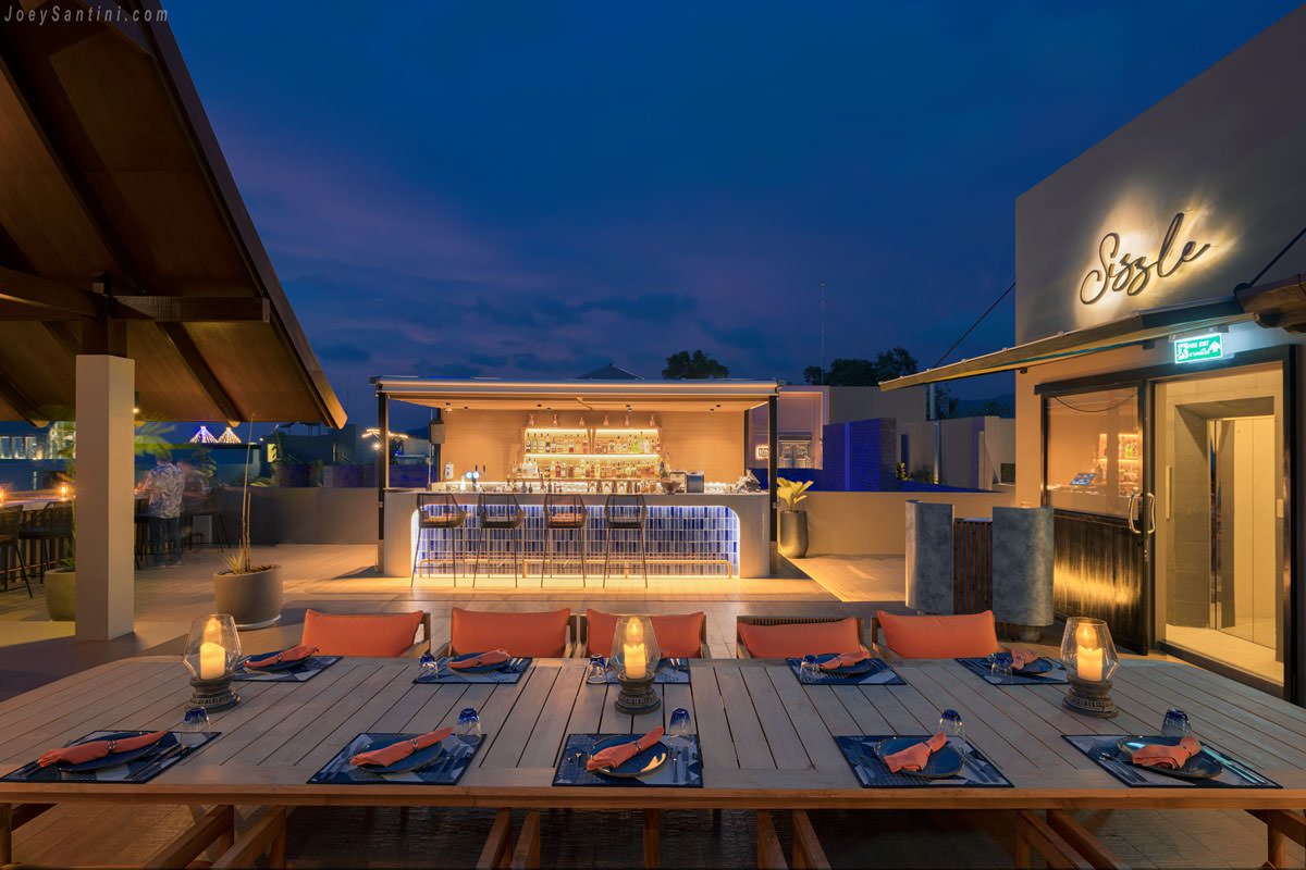 Illuminated cocktail bar and long dining table under a deep blue twilight sky at Sizzle Rooftop Bar.