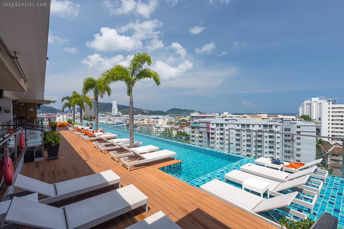 Rooftop infinity pool deck at Skyline Rooftop Marina Phuket with white sun loungers and palm trees under a blue sky.