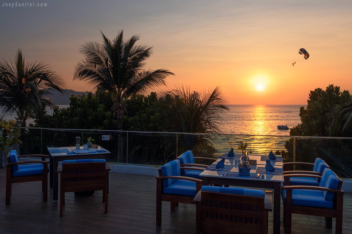 Golden ocean sunset view from The Surface Restaurant and Bar at La Flora Resort with dining tables and a parasailer in the sky.