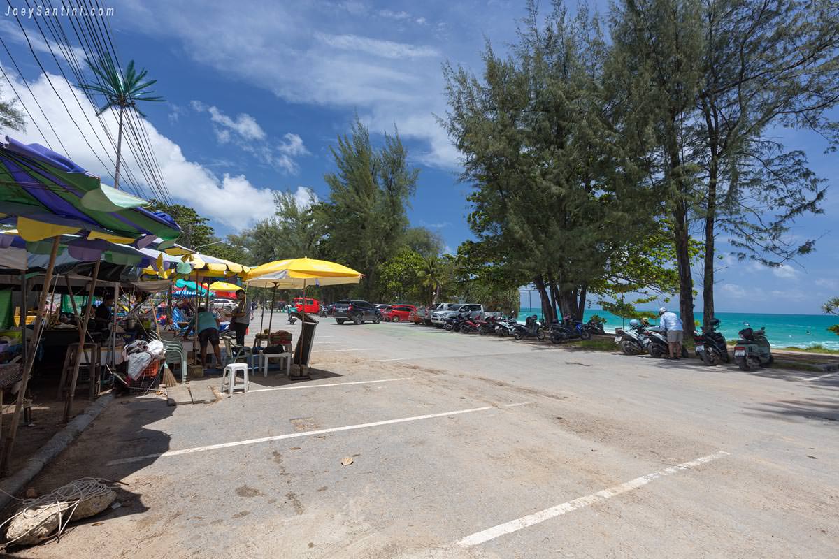 Local food stalls and street vendors with umbrellas next to the beachfront parking area at Surin Beach in Phuket.
