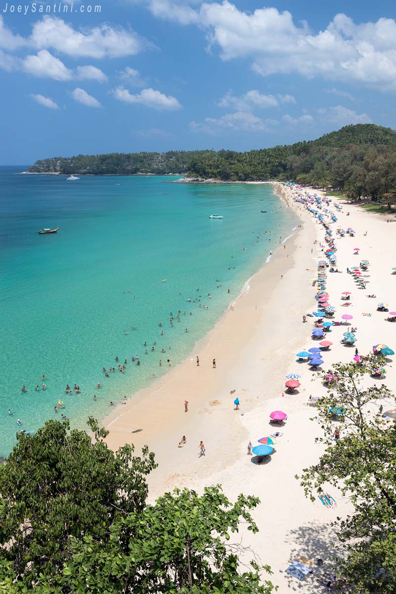 High-angle panoramic view of Surin Beach in Phuket with vibrant turquoise water, white sand, and colorful beach umbrellas.