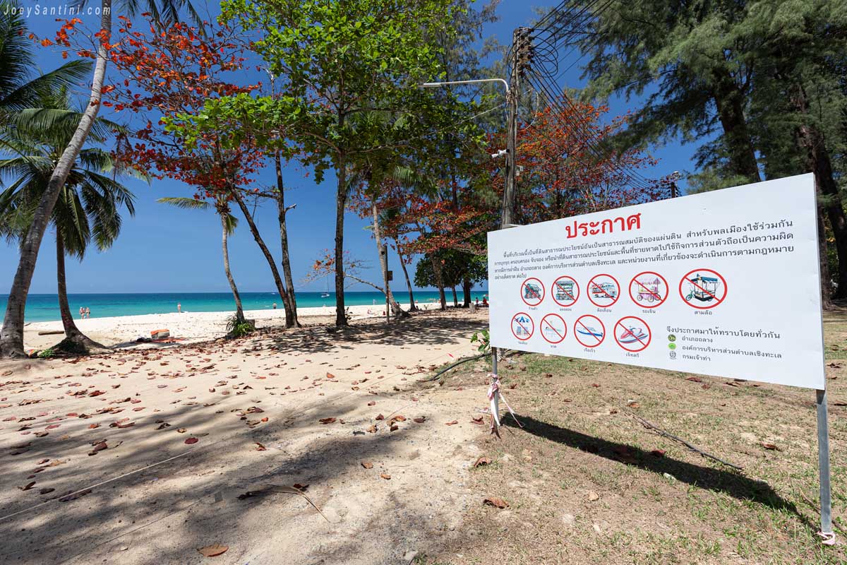An official beach safety and information sign standing on the sand near the trees at Surin Beach.