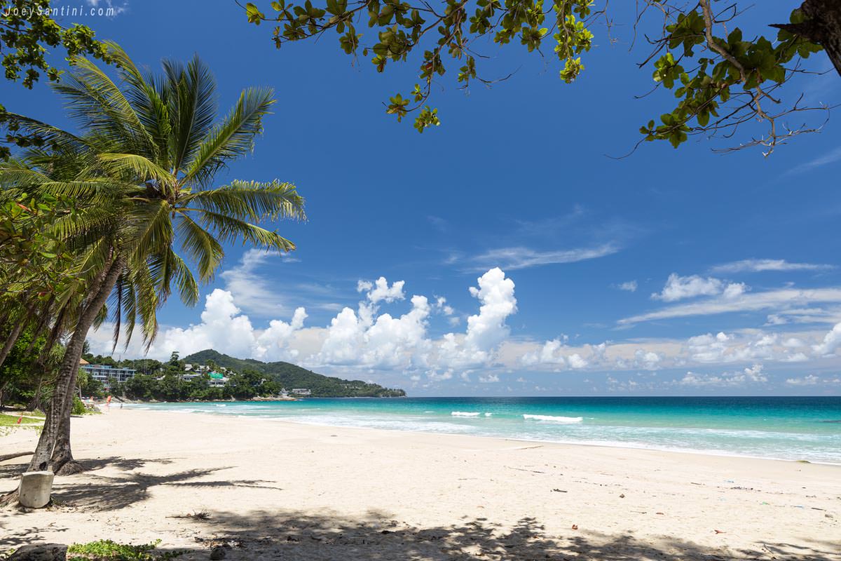 Bright tropical sunshine reflecting on the white sand and calm waves of Surin Beach under scattered white clouds.