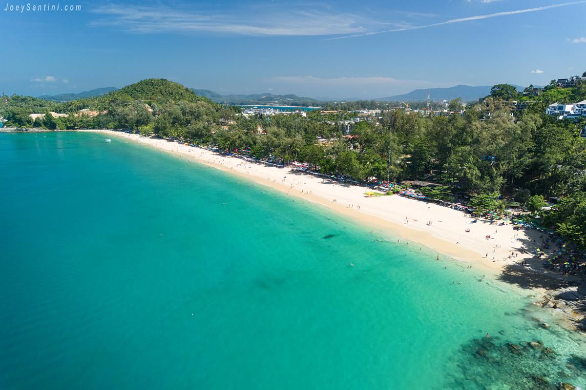 Aerial panoramic view of Surin Beach in Phuket, showing a long stretch of white sand and lush green trees bordering vibrant turquoise ocean water under a clear blue sky.