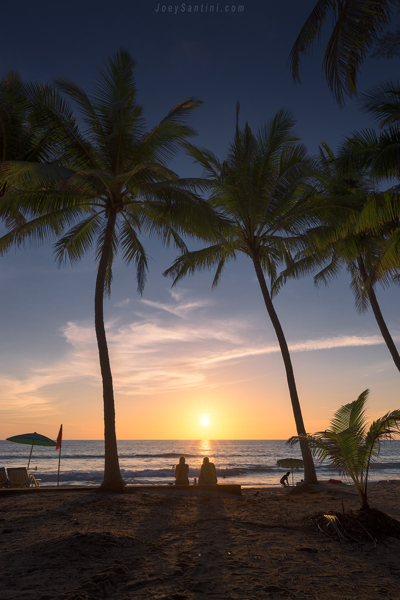 Silhouette of palm trees on Surin Beach at sunset with the low sun on the horizon and warm tropical colors.