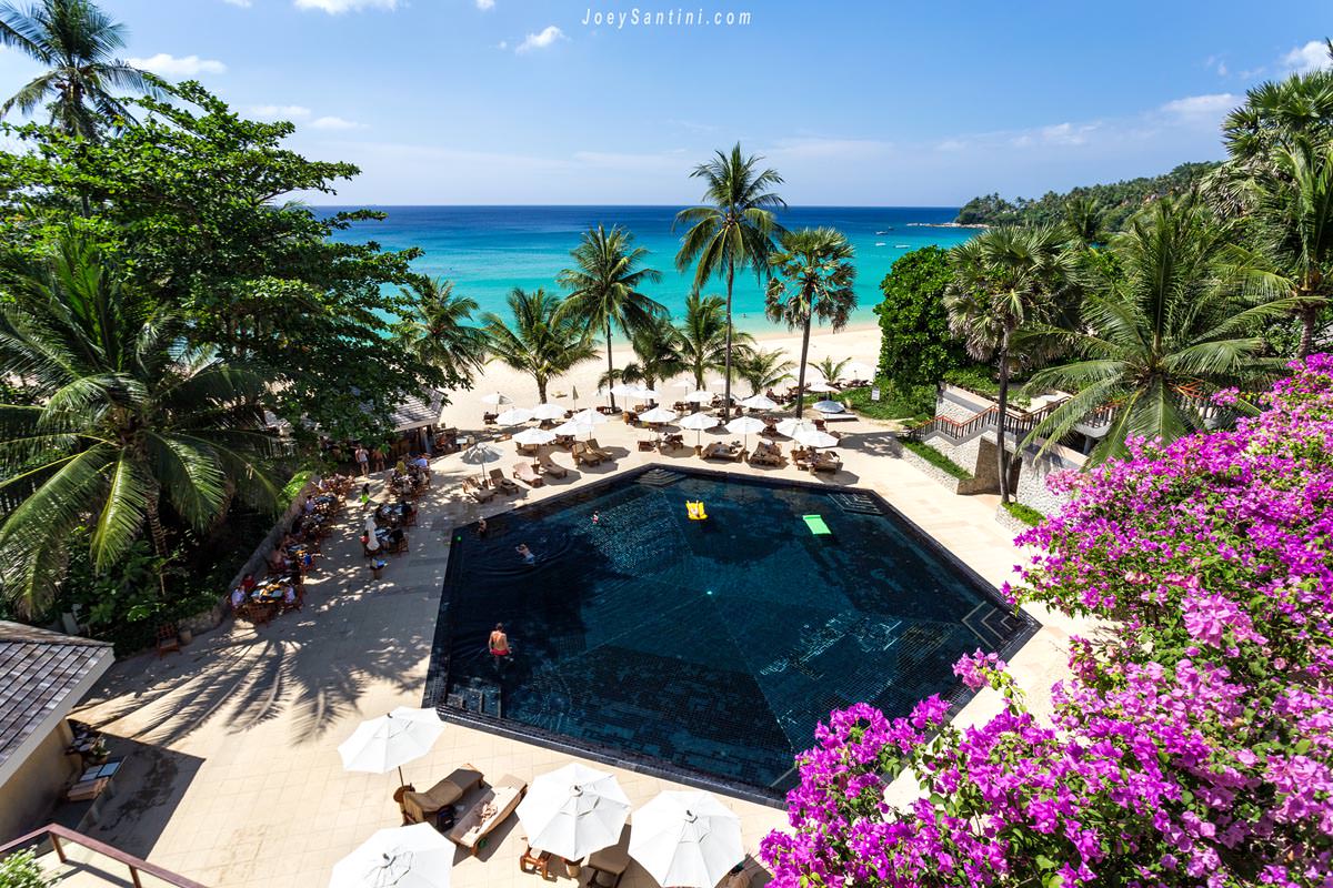 High-angle view of the iconic black-tiled swimming pool at The Surin Phuket resort next to the white sands and turquoise waters of Pansea Beach.