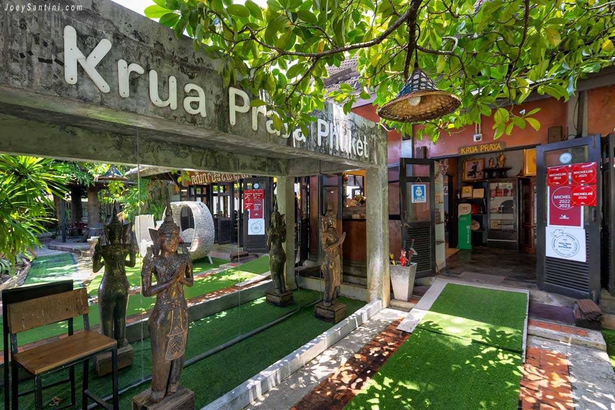 Concrete entrance arch with Krua Praya Phuket name and traditional Thai statues standing on green turf