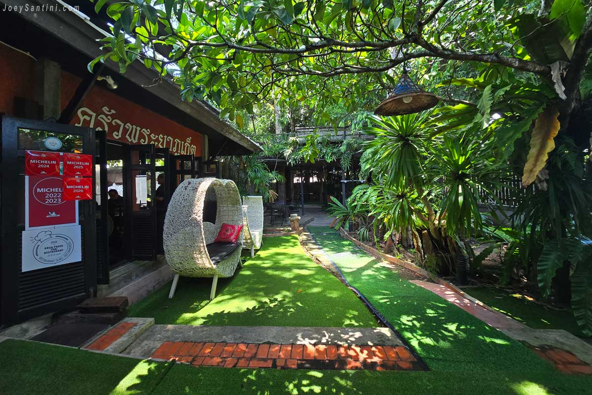 Shaded garden pathway with artificial turf leading to Krua Praya Phuket restaurant entrance with Michelin plaques