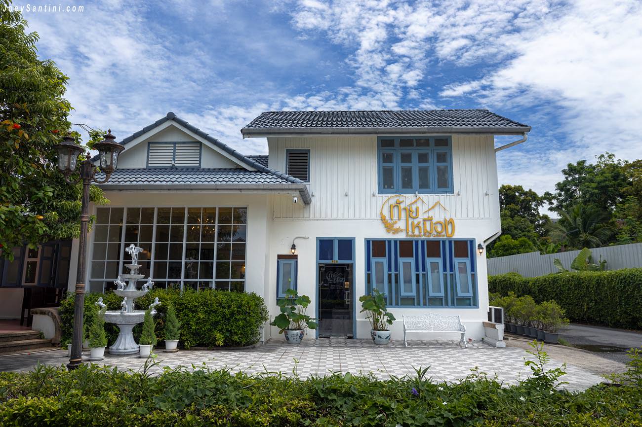 Front view of the two-story white Sino-Portuguese style building with blue window frames, a fountain, and the Thai Mueang Restaurant sign under a blue sky