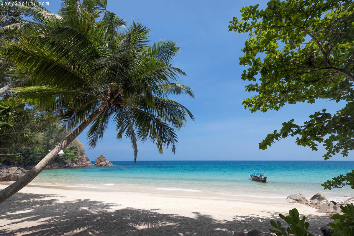A single longtail boat floating in the clear blue sea of Banana Beach Phuket framed by lush tropical foliage