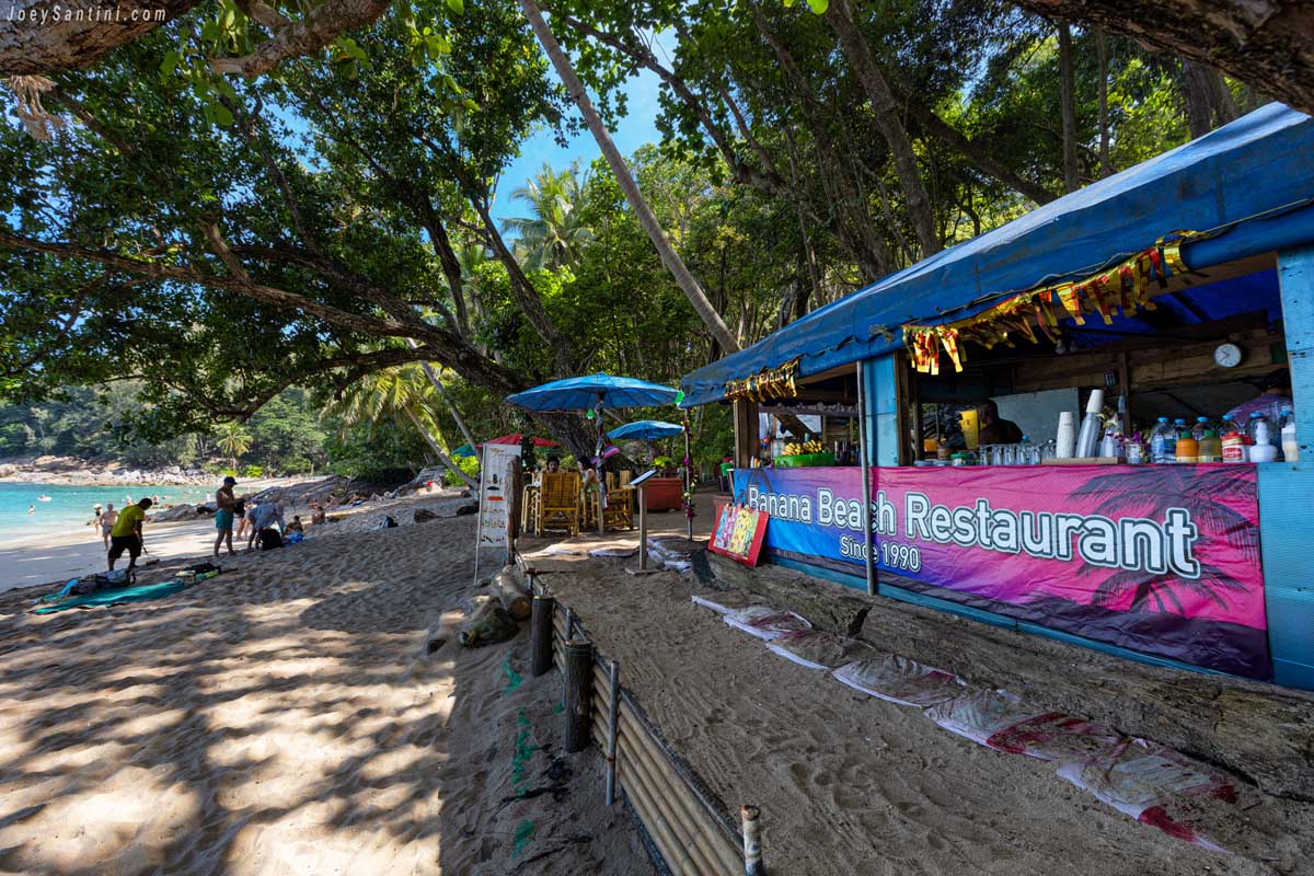 The violet Banana Beach Restaurant sign and shaded dining area under tropical green trees
