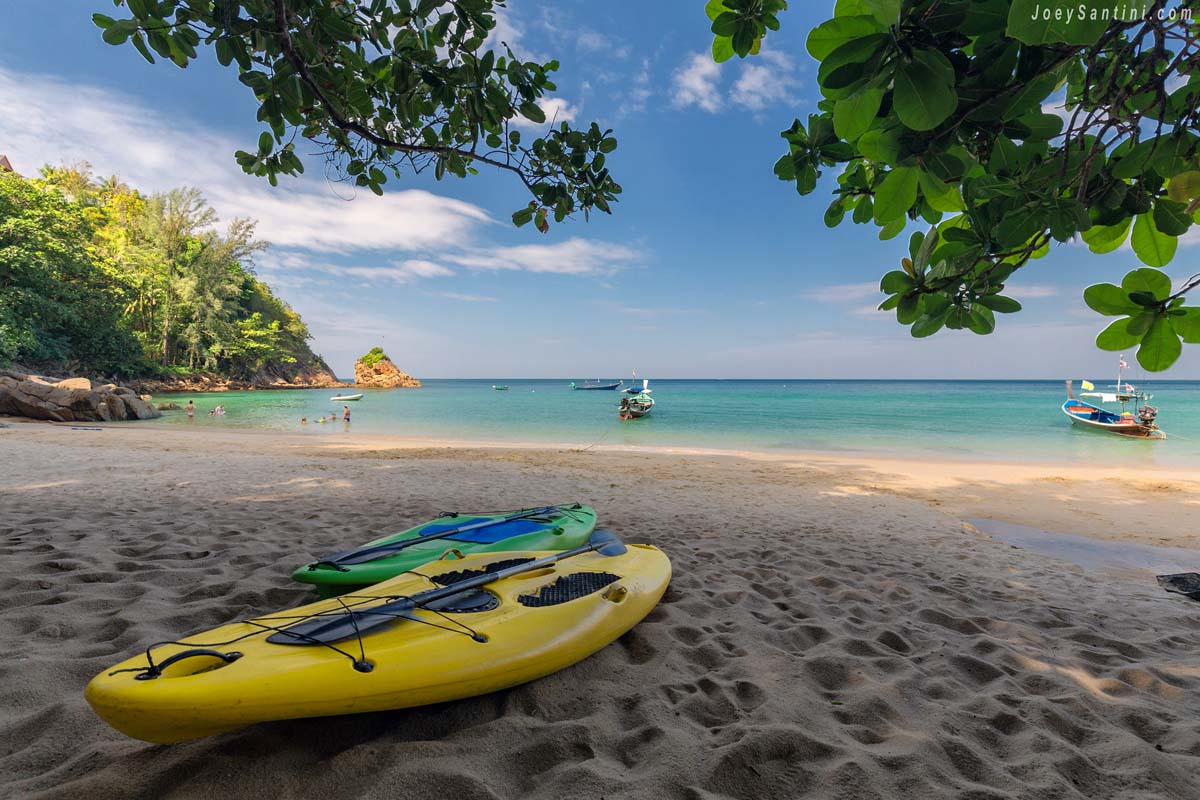Yellow and green rental kayaks resting on the white sand of Banana Beach in Phuket beneath overhanging tree branches